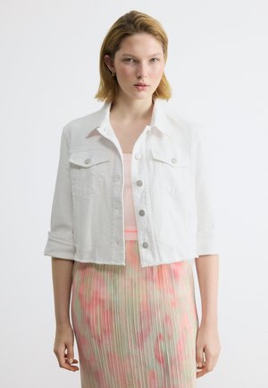 Νεαρή γυναίκα με κοντά ξανθά μαλλιά φοράει ένα λευκό cropped μπουφάν πάνω από ένα παστέλ ροζ τοπ και μια πλισέ φλοράλ φούστα, στέκεται μπροστά σε ένα λευκό φόντο.