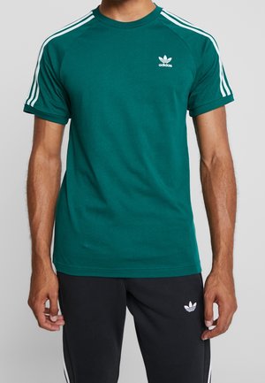 Mann trägt ein grünes Adidas T-Shirt mit weißen Streifen an den Schultern und schwarze Adidas-Hosen mit weißem Logo und seitlichen Streifen.