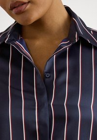 Detailný záber na osobu v tmavomodrej košeli s vertikálnymi bielymi a červenými pruhmi a otvoreným golierom, ktorý odhaľuje časť krku a pier.