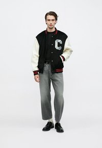 Μαύρο και κρεμ μπουφάν varsity με το γράμμα "C", μανσέτες με κόκκινη και λευκή ρίγα, συνδυασμένο με γκρι τζιν φαρδιάς γραμμής και μαύρα παπούτσια.