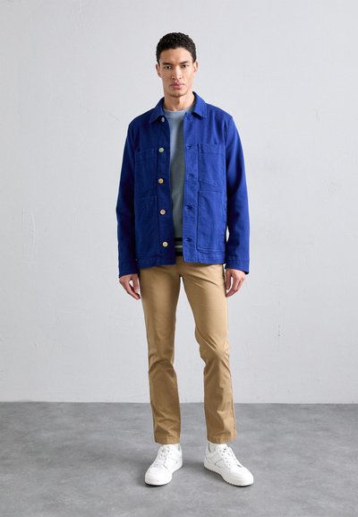 Blue lightweight jacket with four front pockets, paired with a gray shirt, beige pants, and white sneakers. Smooth fabric and button accents.