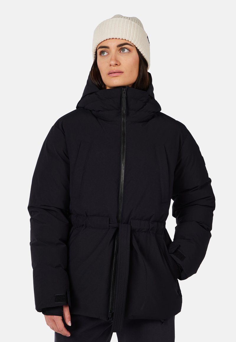 Kvinde iført en sort hættetrøje vinterjakke med frontlynlås og bælte, kombineret med en beige strikket hue, som ser til sin venstre side.