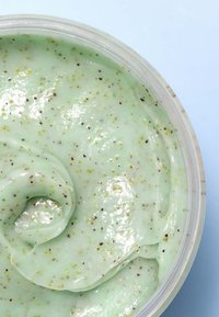 Groene exfolierende scrub met een gladde, glanzende textuur, met fijne donkere en lichte spikkels doorheen, in een doorzichtige container.