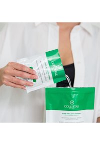 Les enveloppements drainants remodelants Collistar dans un sachet refermable vert et blanc. Une main verse le contenu d'un petit sachet dans le plus grand.