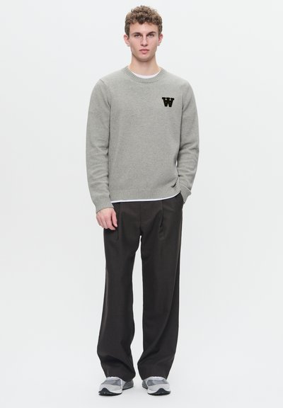 Lys grå strikket sweater med ribbede manchetter, med et sort "W" logo på brystet; parret med løs siddende brune bukser og grå sneakers.
