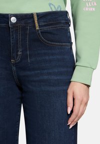 Granatowe dżinsy z złotymi stębnowaniami, pięcioma kieszeniami i zapięciem na zamek z przodu. W połączeniu z jasnoskalowym topem z długim rękawem.