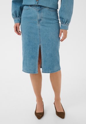 Femme portant une jupe en jean bleue longueur genou avec une fente à l’avant et des talons pointus marron, debout devant un fond uni.