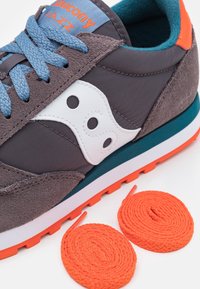 Saucony Jazz -tennarit, joissa on harmaa tekstiili- ja mokkanahkainen päällinen, valkoinen logo, siniset nauhat, teal-väriset yksityiskohdat ja oranssi kumipohja, jossa on kaksi rullattua oranssia nauhaa.
