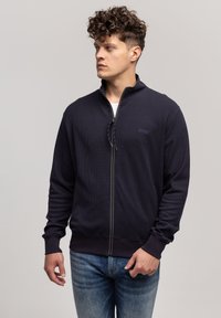 Granatowa kurtka zapinana na zamek o fakturowanej tkaninie, z kołnierzem stójkowym i ściągaczami. Posiada małe logo na klatce piersiowej oraz zamek błyskawiczny na całej długości.