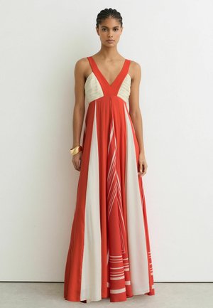 Femme portant une robe longue rouge et blanche sans manches avec un profond décolleté en V et un bracelet en or, debout contre un mur blanc uni.