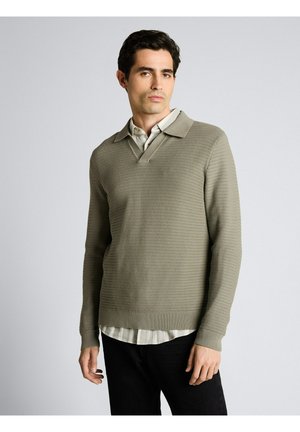 Un homme se tient face à l'avant, portant un pull en maille beige par-dessus une chemise à col rayée et un pantalon noir, sur un fond uni.