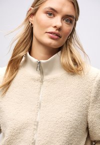 Beżowa kurtka polarowa z wysokim kołnierzem i zapięciem na zamek. Teksturowany materiał o miękkim wyglądzie. Modelka ma blond włosy i delikatny makijaż.