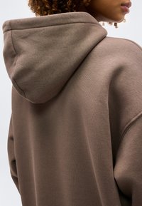 Άτομο με σγουρά μαλλιά που φοράει μια φαρδιά καφέ μπλούζα με κουκούλα, εμφανιζόμενο από πίσω και πλάγια, με την κουκούλα σηκωμένη.