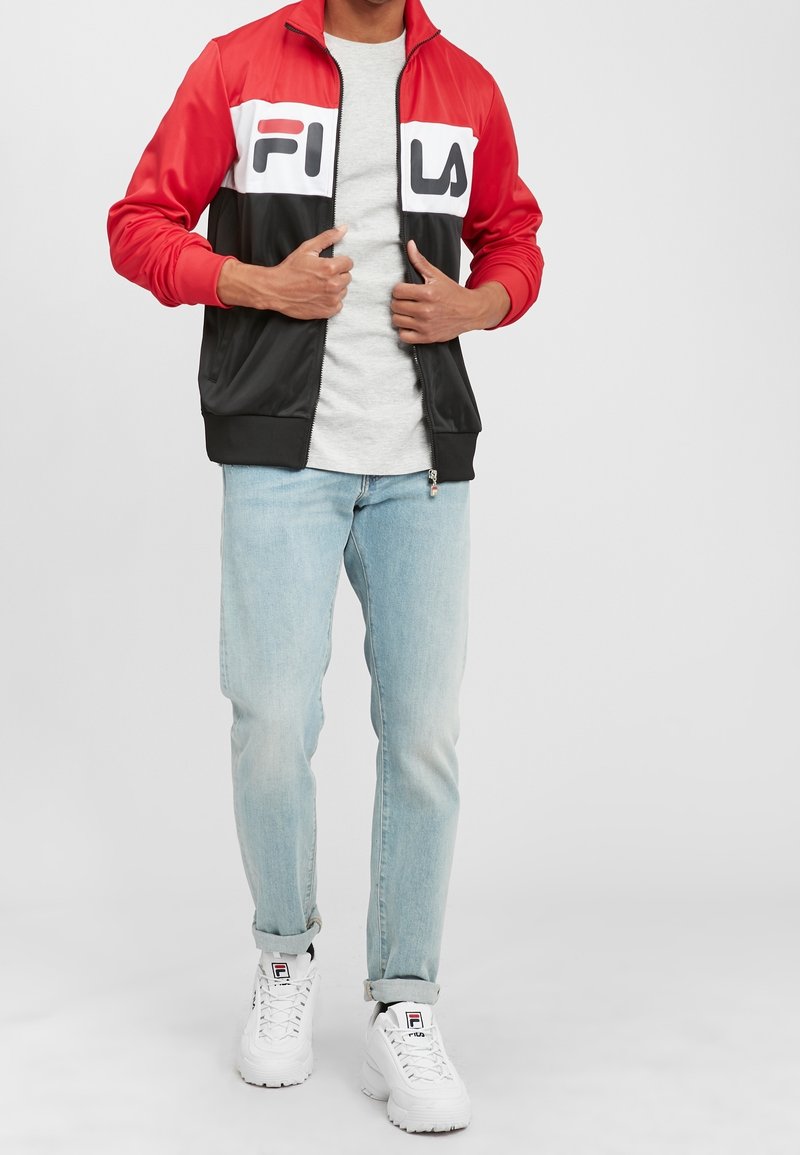 Homme portant une veste FILA rouge, blanche et noire, un t-shirt gris clair, un jean bleu clair et des baskets blanches FILA, debout sur un fond blanc.