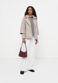Manteau en mélange de laine beige avec des manches oversized, haut noir et blanc rayé, pantalon large blanc, sac bordeaux avec un nœud, et chaussures noires.