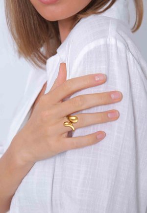 Guldbelagt metalring med et snoet design, båret på en hånd hvile på en hvid tekstureret stofskjorte. Korte, naturlige negle kan ses.