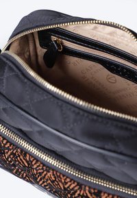 Interior de una bolsa de tela acolchada negra que muestra un bolsillo con cremallera, forro beige con texto de la marca y un acento texturizado marrón a lo largo de la base. Detalle de cremallera dorada.