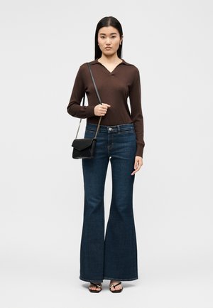 Femeie purtând un tricou polo maro cu mâneci lungi, blugi evazați de culoare albastru închis, sandale negre și o geantă neagră de umăr, pe un fundal simplu.