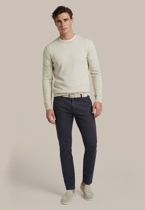 Man draagt een beige trui, donkere smalle broek, lichte geweven riem en beige instapschoenen, staand tegen een effen achtergrond.