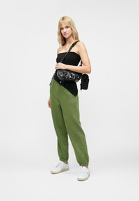 Black fitted tank top, green tapered sweatpants, white sneakers, and a black shoulder bag. Simple design and smooth textures throughout.
