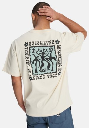 Mężczyzna w beżowej koszulce Quiksilver z czarnymi abstrakcyjnymi figurami i napisem „The Original Quiksilver Boardriding Co. Since 1969” na plecach, ręka na głowie.