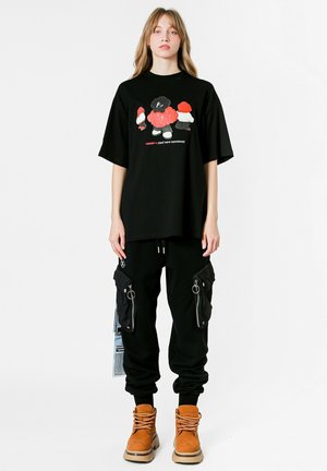 Camiseta gráfica negra con diseño de dibujos animados colorido, combinada con pantalones cargo negros con bolsillos con cremallera, y botas de cordones marrones.