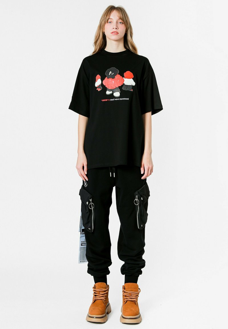 Camiseta gráfica negra con diseño de dibujos animados colorido, combinada con pantalones cargo negros con bolsillos con cremallera, y botas de cordones marrones.