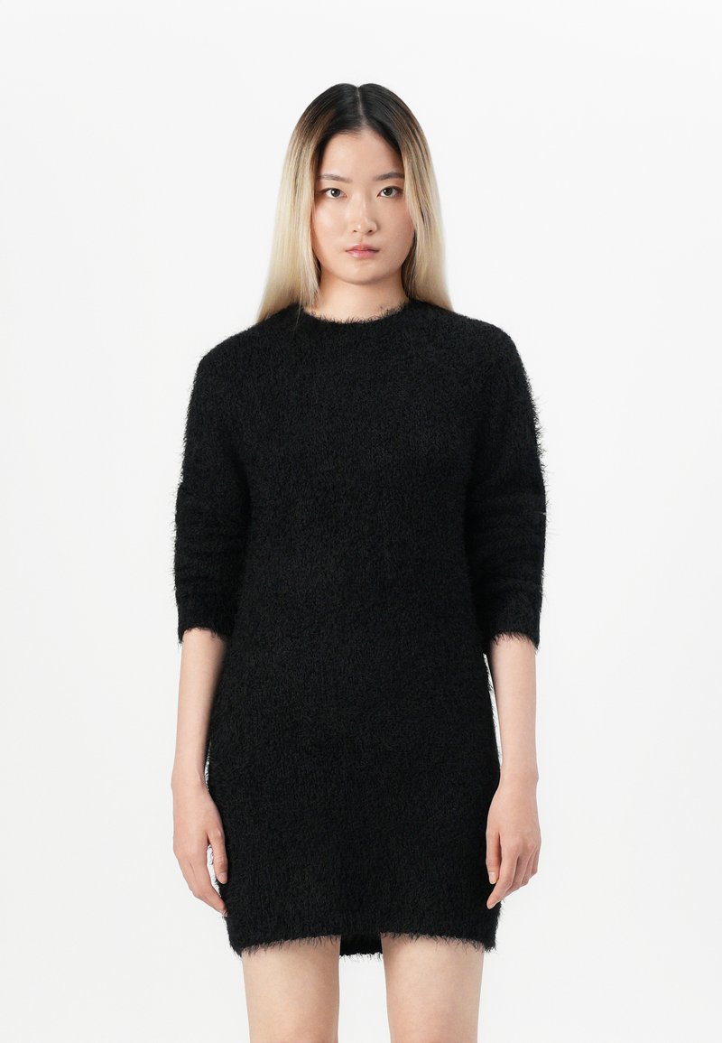 Junge Frau mit langen glatten Haaren, die ein schwarzes, flauschiges Strickkleid mit Dreiviertelärmeln trägt und vor einem schlichten weißen Hintergrund steht.