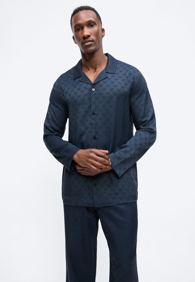 Férfi sötétkék, hosszú ujjú gombos ingben és hozzáillő, finom textúrájú mintás nadrágban, egyszínű fehér háttér előtt áll.