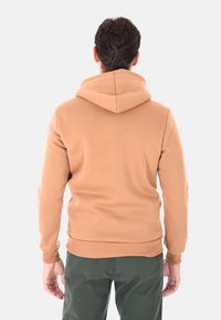 Uomo che indossa un hoodie beige e pantaloni verde scuro, in piedi rivolto di spalle contro uno sfondo bianco semplice.