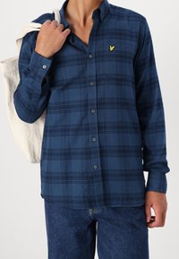Granatowa koszula w kratę zapinana na guziki, wykonana z tkaniny o miękkiej fakturze; posiada kieszeń z złotym logo oraz długie rękawy. Model trzyma płócienną torbę na ramię.