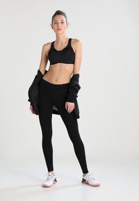 Soutien-gorge de sport noir et leggings, design ajusté, texture lisse, superposés avec une veste noire, associés à des chaussures de sport blanches et roses.