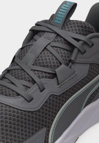 Primo piano di una sneaker grigia in mesh con lacci grigi, logo Puma turchese sulla linguetta e suola bianca con dettagli turchesi.