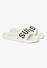 Hvide slidesandaler med en bred rem, der har fed sort "SUPERDRY" skrift. Glat materiale; flad sål med diskret branding på siden.
