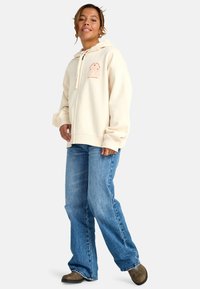 Felpa crema con zip frontale e coulisse, caratterizzata da un design stampato sul petto. Abbinata a jeans blu larghi e scarpe slip-on scure.