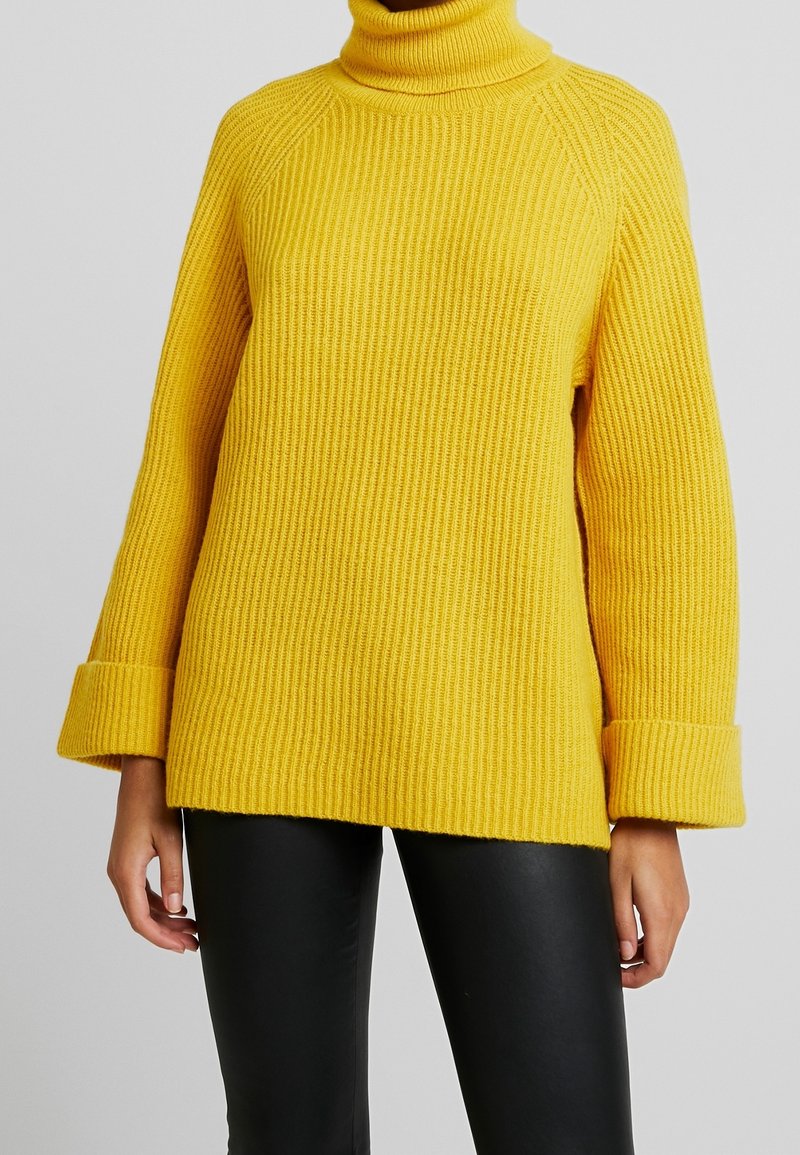 Femme portant un pull à col roulé en maille côtelée jaune vif avec de larges poignets et un pantalon noir ajusté, sur un fond uni.