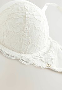 Witte kantenslips met ingewikkelde bloemmotieven, een textuursteek en gouden accenten op de band.