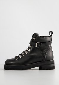 Bottine en cuir noir avec œillets métalliques argentés, semelle épaisse, bride à la cheville avec boucle, et languette à l'arrière.