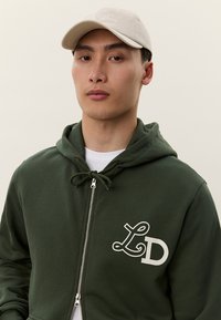 Mladá osoba nosí béžovú čiapku a tmavozelenú mikinu na zips s viazacou šnúrkou a logom "LD" na hrudi, stojac pred jednofarebným pozadím.