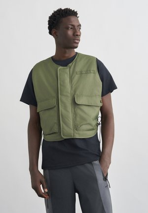 Férfi, aki fekete póló fölött olívazöld mellényt visel első zsebekkel és szürke nadrágot, oldalra néz, egyszínű háttér előtt.