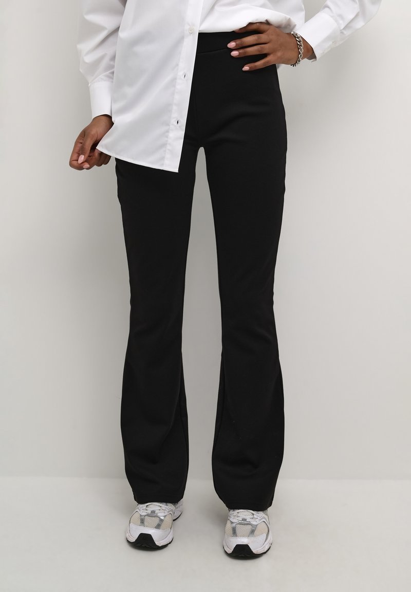 Pantalons évasés noirs en matériau extensible, avec une texture lisse et une taille ajustée, associés à une chemise blanche à boutons et des baskets.