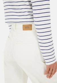 Weiße Jeans mit geradem Bein, versehen mit einem hellbraunen Lederlabel auf der Gesäßtasche. Dazu ein gestreiftes Langarmshirt.