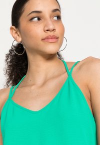 Top verde sem mangas com alças finas, apresentando um decote em V, textura suave e design minimalista. Brincos de argola prateados em forma circular visíveis.