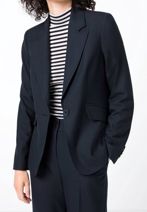 Person trägt einen schwarzen, maßgeschneiderten Blazer und passende Hose über einem schwarz-weiß gestreiften Rollkragentop und steht vor einem weißen Hintergrund.