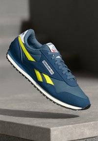 Mėlynos ir geltonos Reebok sportbačiai, turintys lygaus odos ir zomšo viršutinę dalį, plokščius raiščius ir tekstūruotą guminį padą su logotipo akcentais.