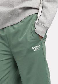 Zaļas sporta bikses ar gludu tekstūru, ar sānu kabatām un baltu Reebok logo kreisajā apakšējā kājā.