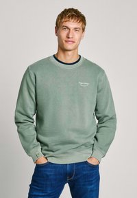Grön sweatshirt med rund halsringning, långa ärmar med mudd och en liten logotyp på bröstet. Kombinerad med blå jeans. Mjuk, strukturerad tyg.