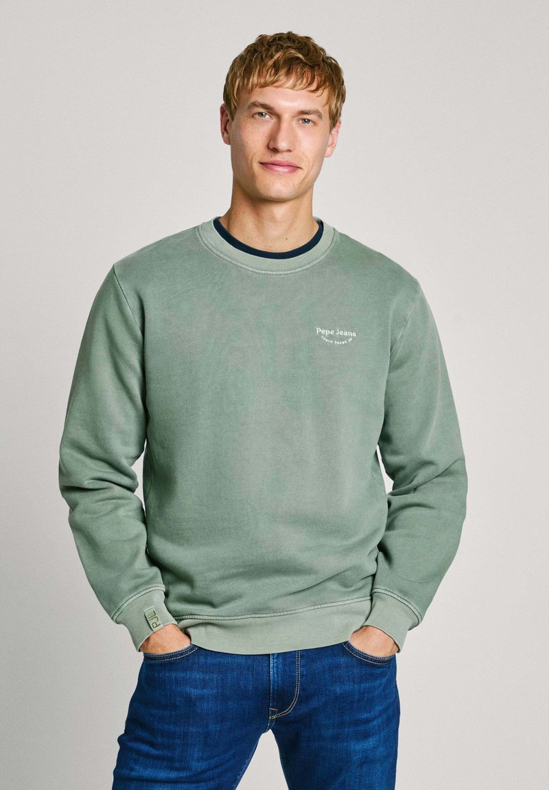 Grön sweatshirt med rund halsringning, långa ärmar med mudd och en liten logotyp på bröstet. Kombinerad med blå jeans. Mjuk, strukturerad tyg.