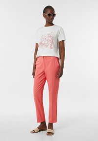 Witte t-shirt met een rode kreeftprint, gecombineerd met koraalkleurige op maat gemaakte pantalons. Het model draagt ronde zonnebrillen en platte geweven slidesandalen.