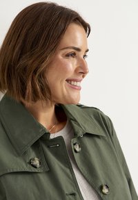 Olijfgroen trenchcoat met grote knopen, een opvallende kraag en een gladde stofstructuur, vergezeld van een lichte, textuurrijke trui eronder.